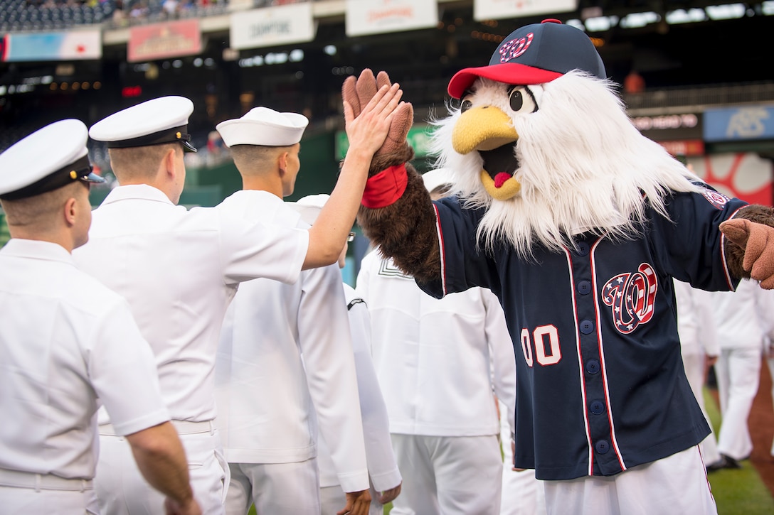 Sailors line up and high-five a mascot in a baseball stadium.