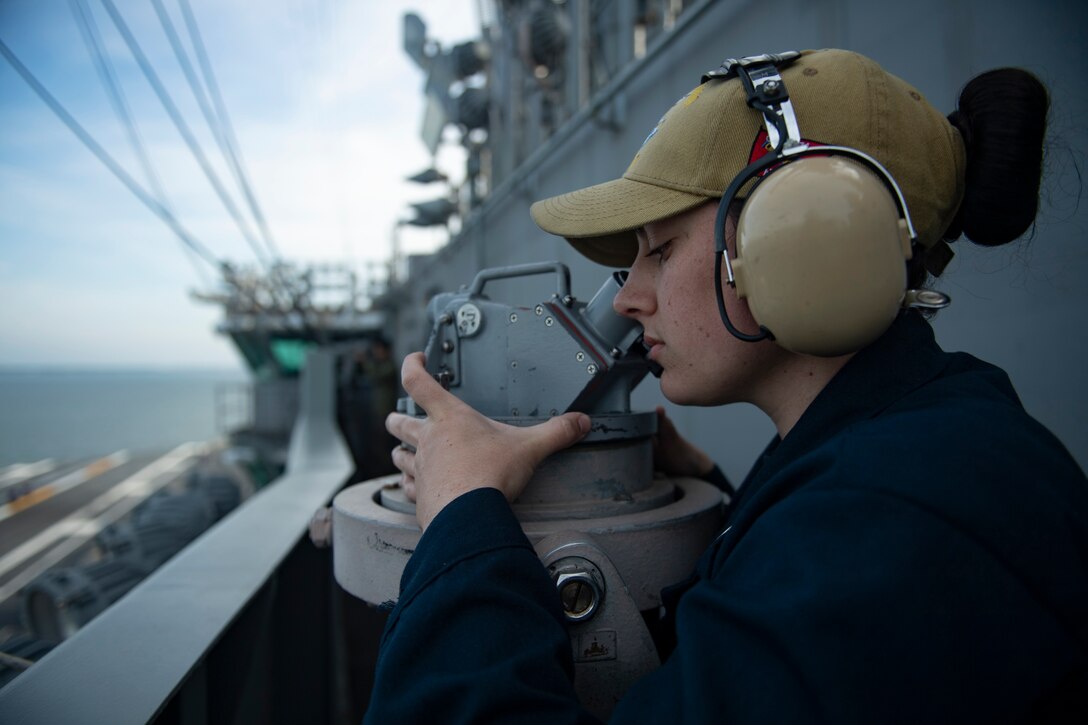 A sailor wearing a cap and headphones looks into a sextant aboard a ship.