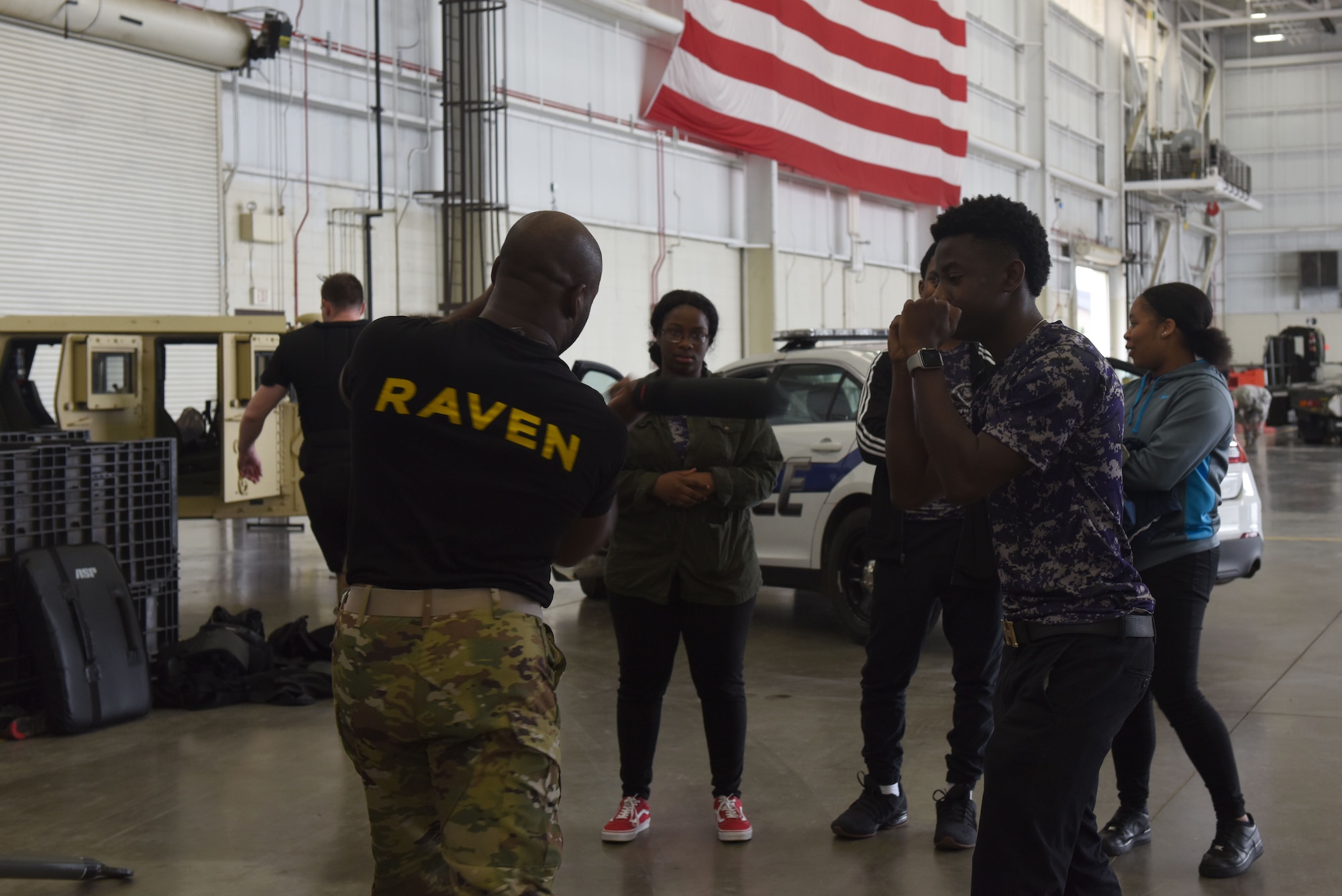 A man wearing a black shirt saying "RAVEN" on his back says stands next to a high school student with his fists up in a sparring position.