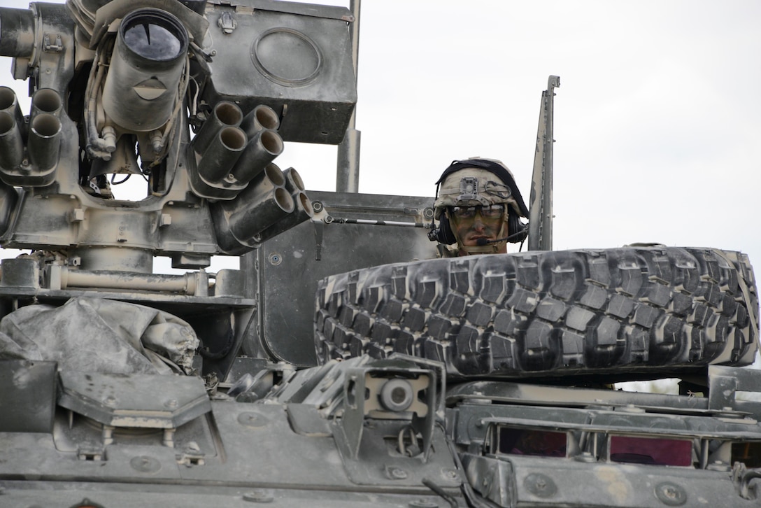 A soldier’s camouflaged face emerges from the hatch of an armored vehicle above a spare tire.