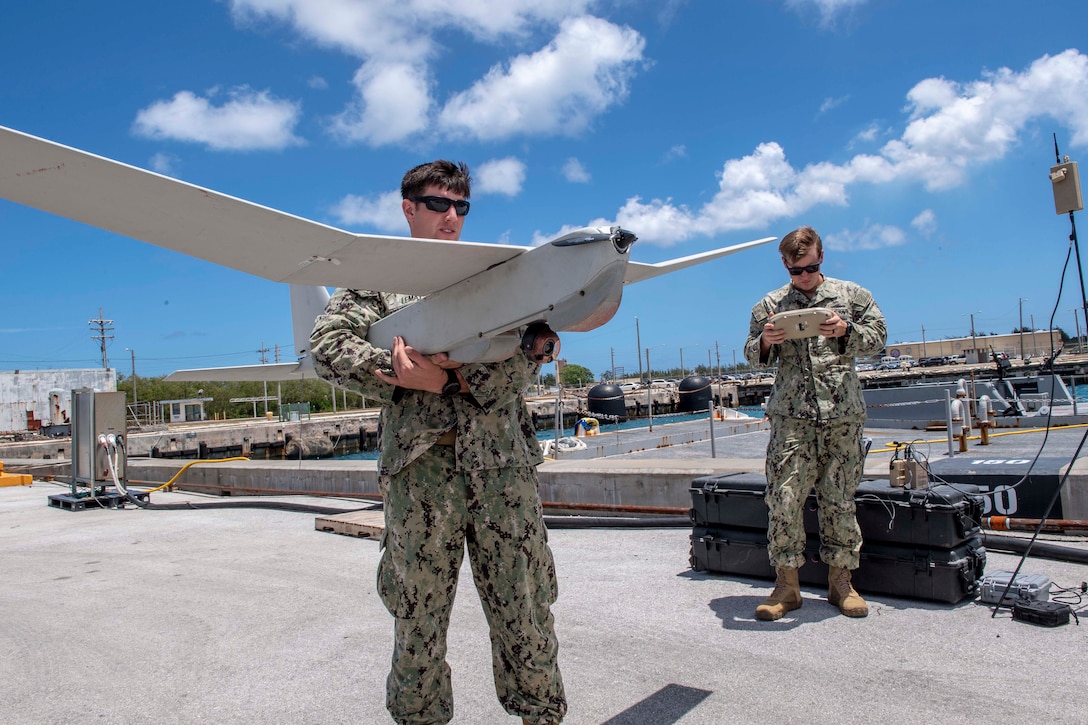 A sailor wearing sunglasses holds a winged drone under his arm while another sailor checks a remote control.