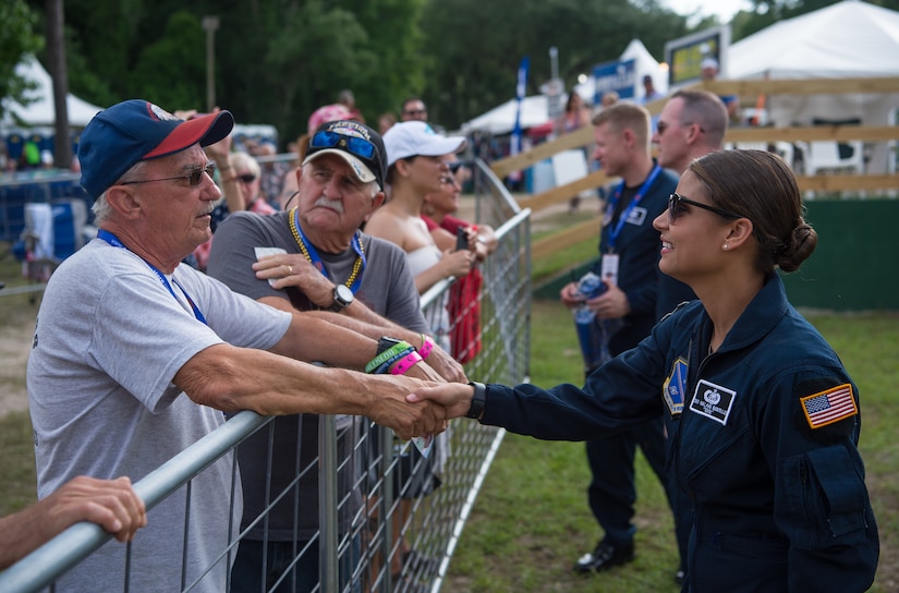 A female airman shakes hands with an older man on the opposite side of a fence. Others can be seen doing the same in the background at a fair.