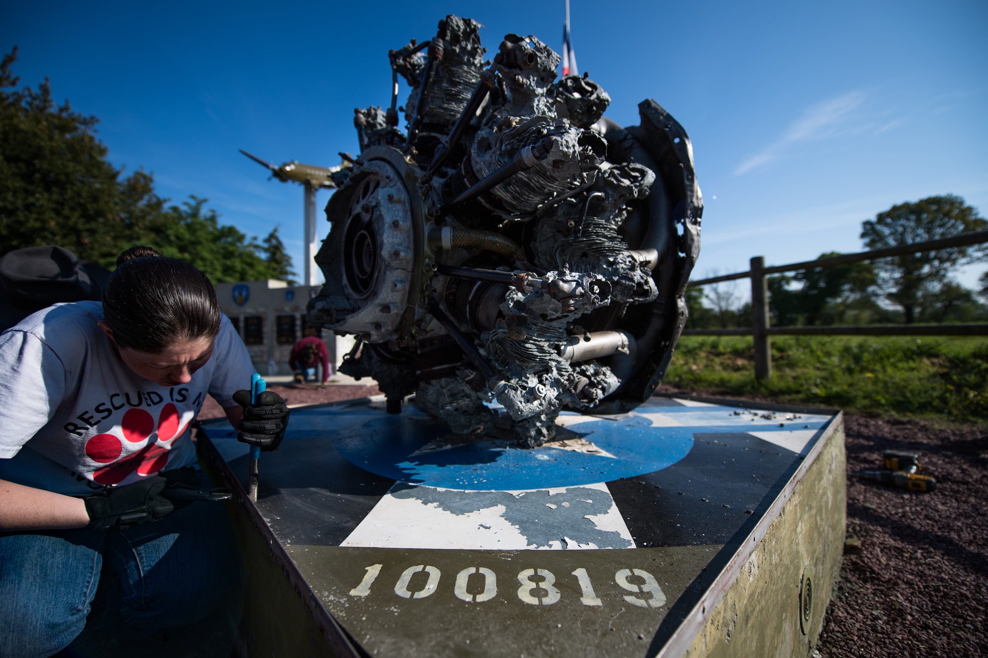 U.S. Air Force Airman 1st Class Rachel Miller, 435th Construction Training Squadron power production journeyman, loosens a screw attached to the pedestal of a WWII aircraft display at a memorial in Picauville, France, April 30, 2019. The “100819” painted on the pedestal represents the aircraft number the engine belonged to. (U.S. Air Force photo by Staff Sgt. Devin Boyer)