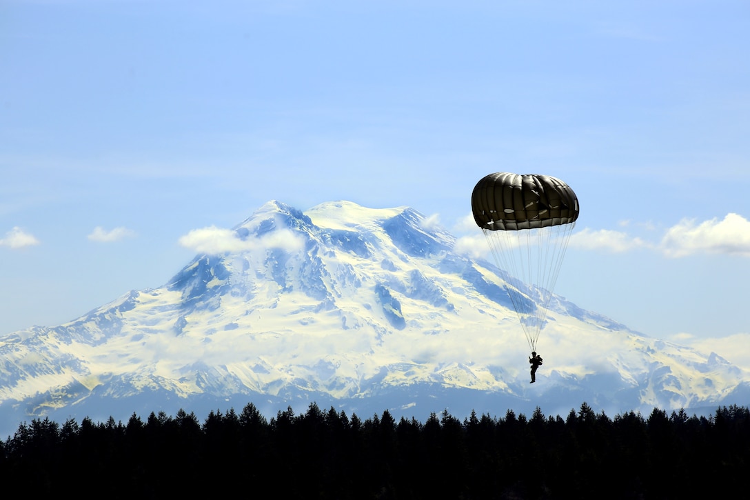 A parachuting soldier glides above the trees as a massive mountain looms in the background.