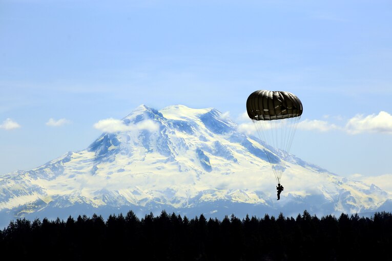 A parachuting soldier glides above the trees as a massive mountain looms in the background.