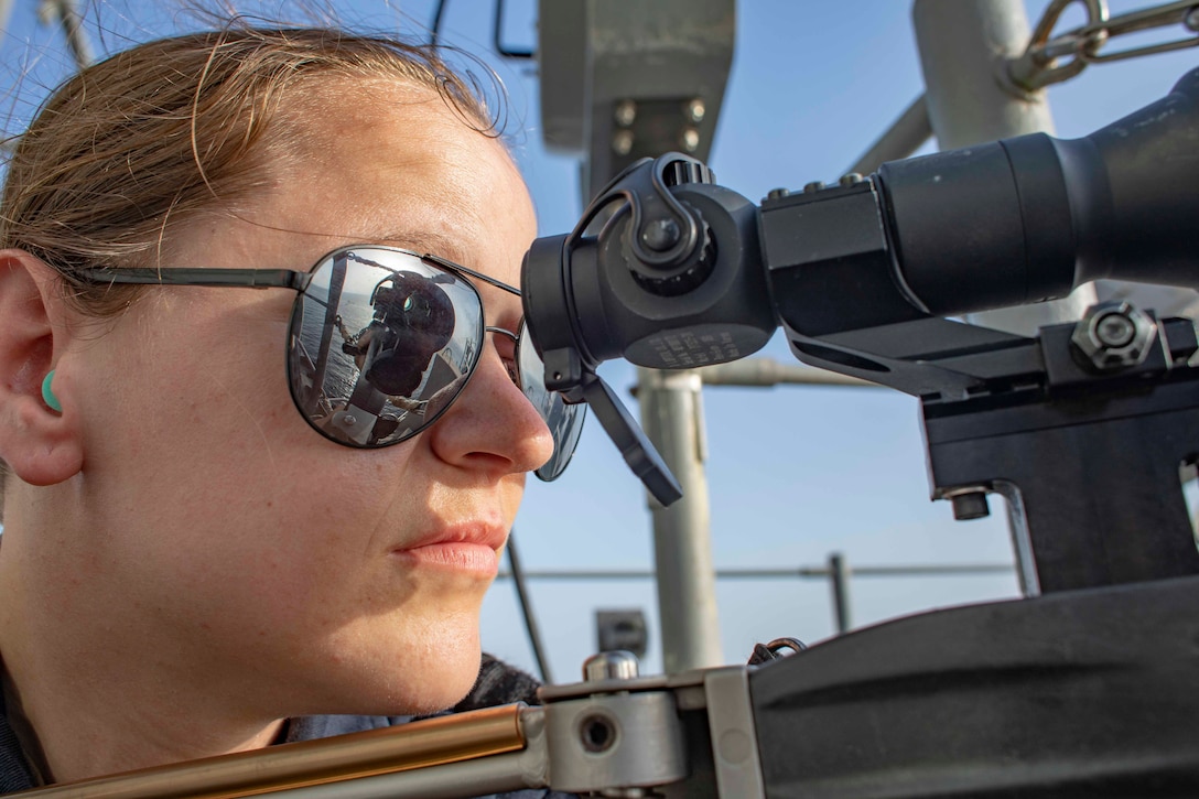 A sailor looks through a telescope as the water ahead is reflected in sunglasses.