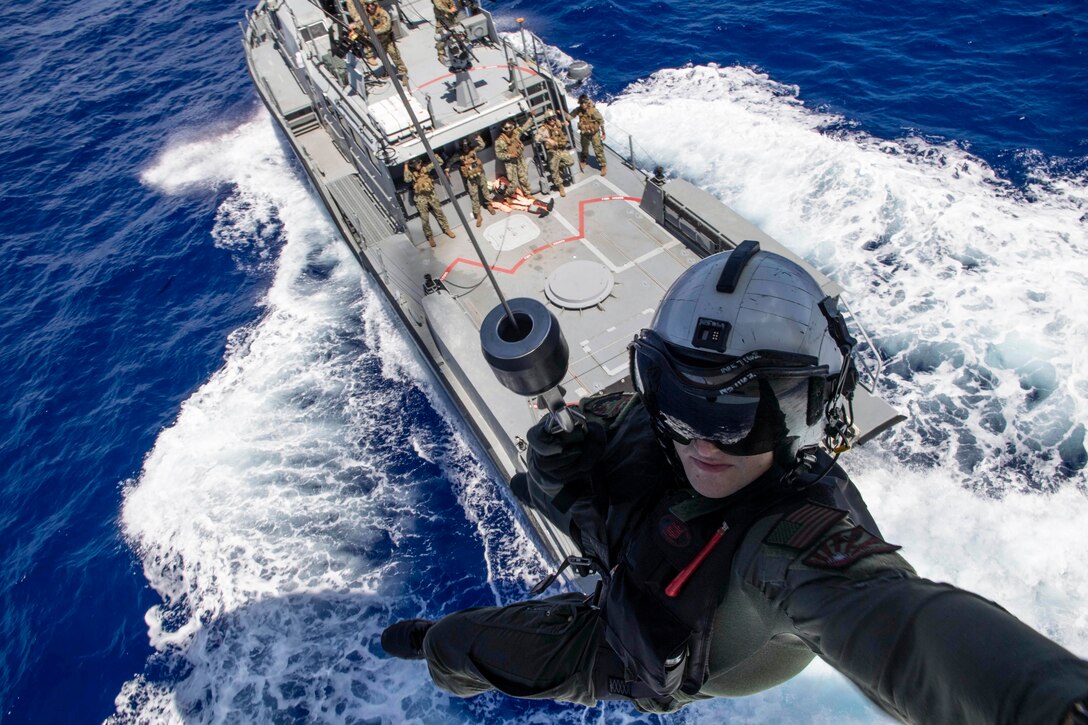 A sailor wearing a harness hangs out of a helicopter above a boat with people on standing on the deck.