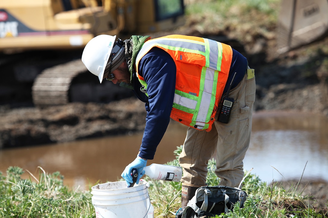 A man in a construction vest and hard hat sprays liquid into a bucket in front of standing water.