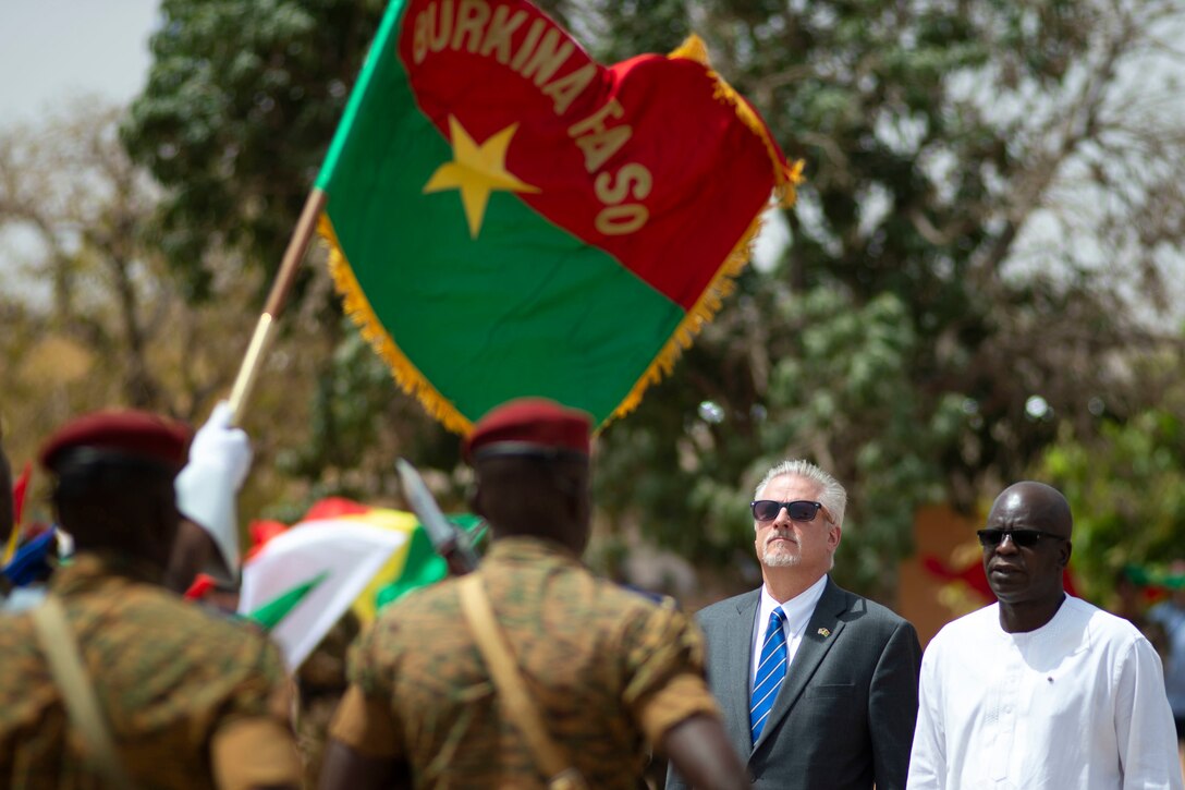 Soldiers march with a flag as civilian men observe.