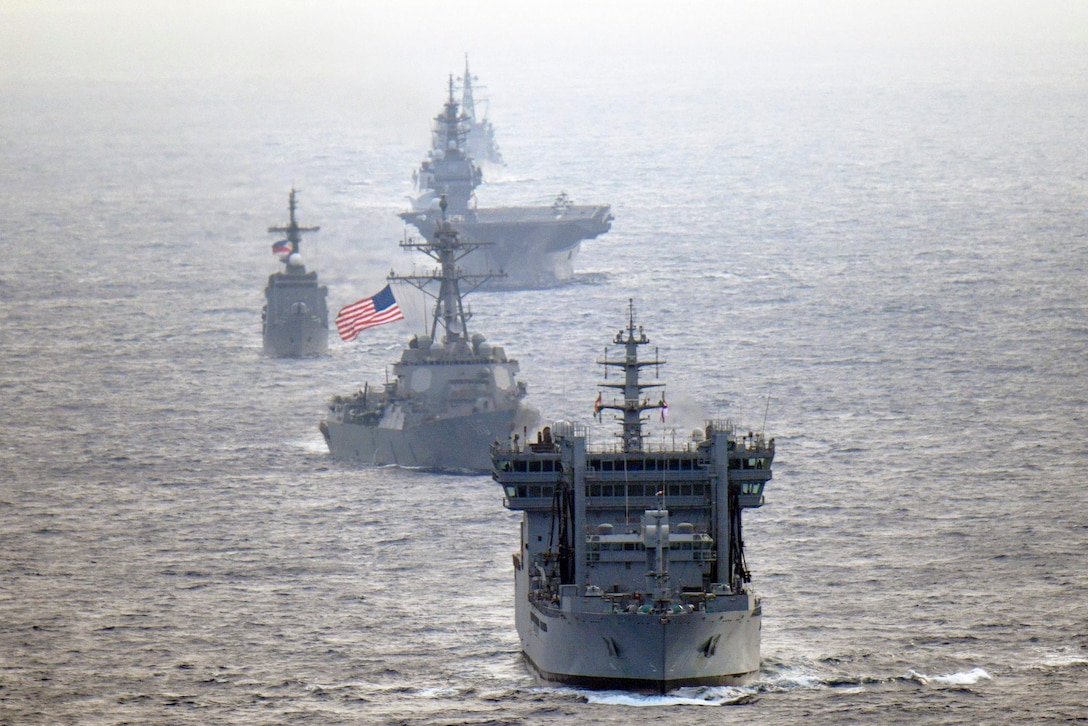 Six ships, with one displaying an American flag, move through water.