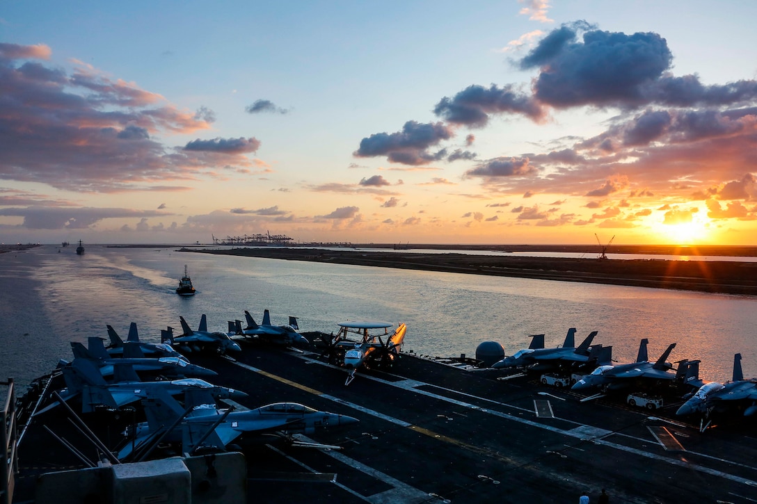 An aircraft carrier transits a canal while the sun sets.