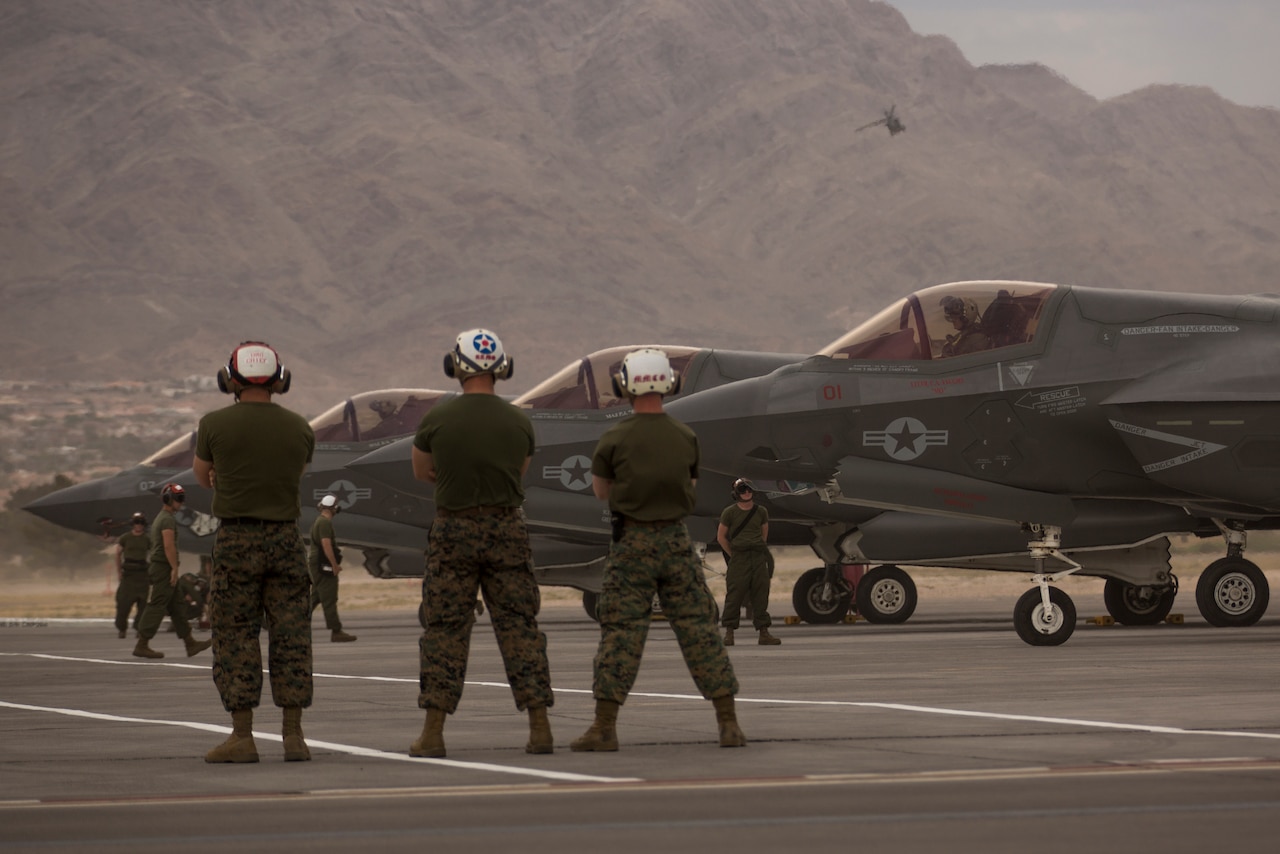 Marines stand with their arms folded as pilots prepare their aircraft for takeoff.