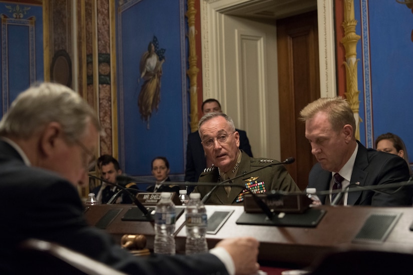 A Marine in uniform and a civilian sit behind microphones at a table.