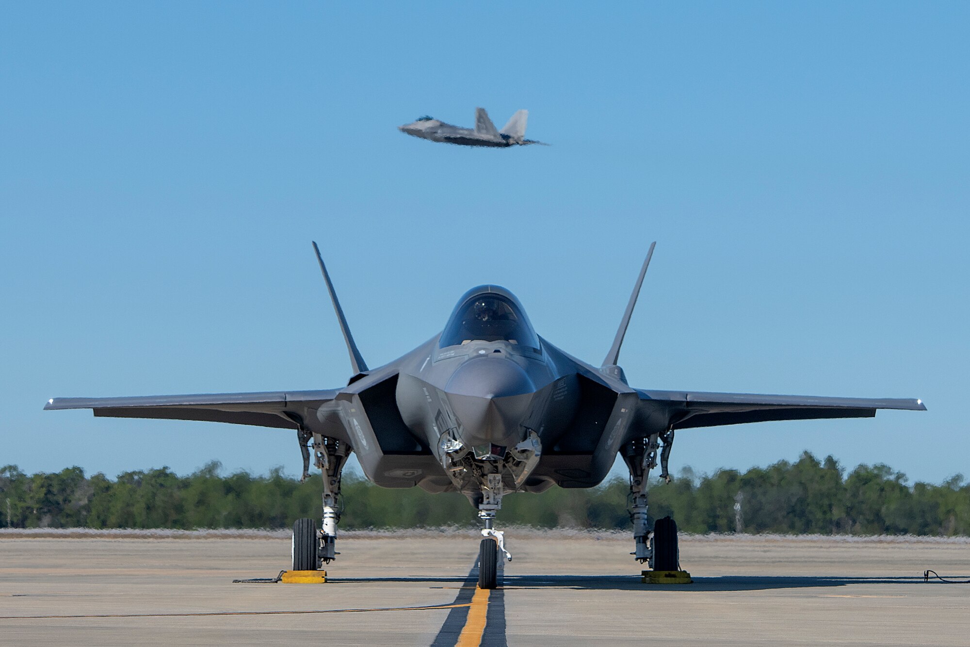 An F-35A Lightning II assigned to the 58th Fighter Squadron awaits permission to taxi as an F-22 Raptor assigned to the 27th Fighter Squadron takes off in the background, Dec. 4, 2018, at Eglin Air Force Base, Fla. (U.S. Air Force Staff Sgt. Peter Thompson)
