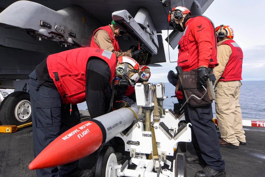 Five sailors wearing red vests, helmets, earphones and safety goggles stand beneath an aircraft and secure a missile.
