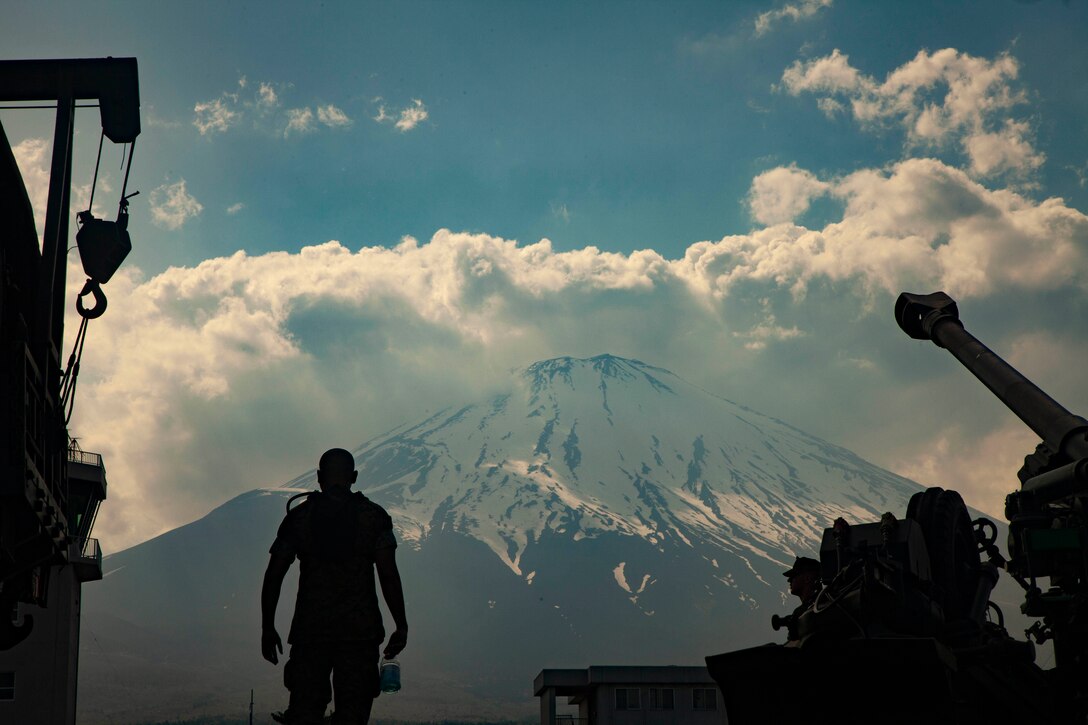 A Marine holding a sports drink bottle stands between a guard tower and an armored vehicle in front of a massive snowcapped mountain.