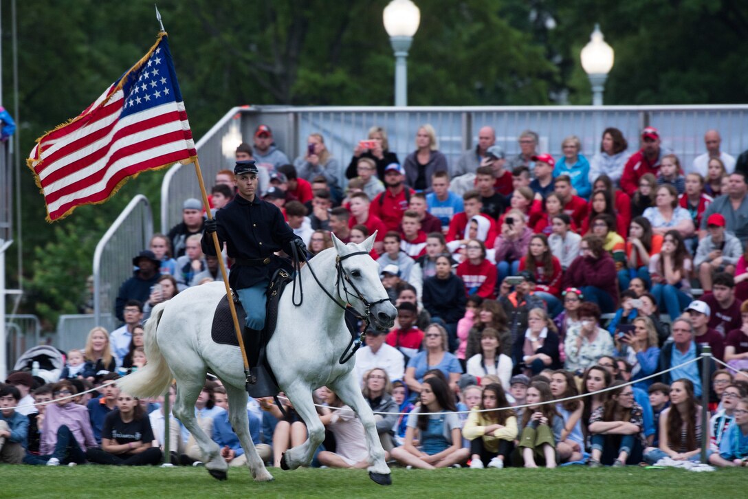 A soldier wearing a vintage uniform and cover holds a flag while riding a white horse in front of a crowd.