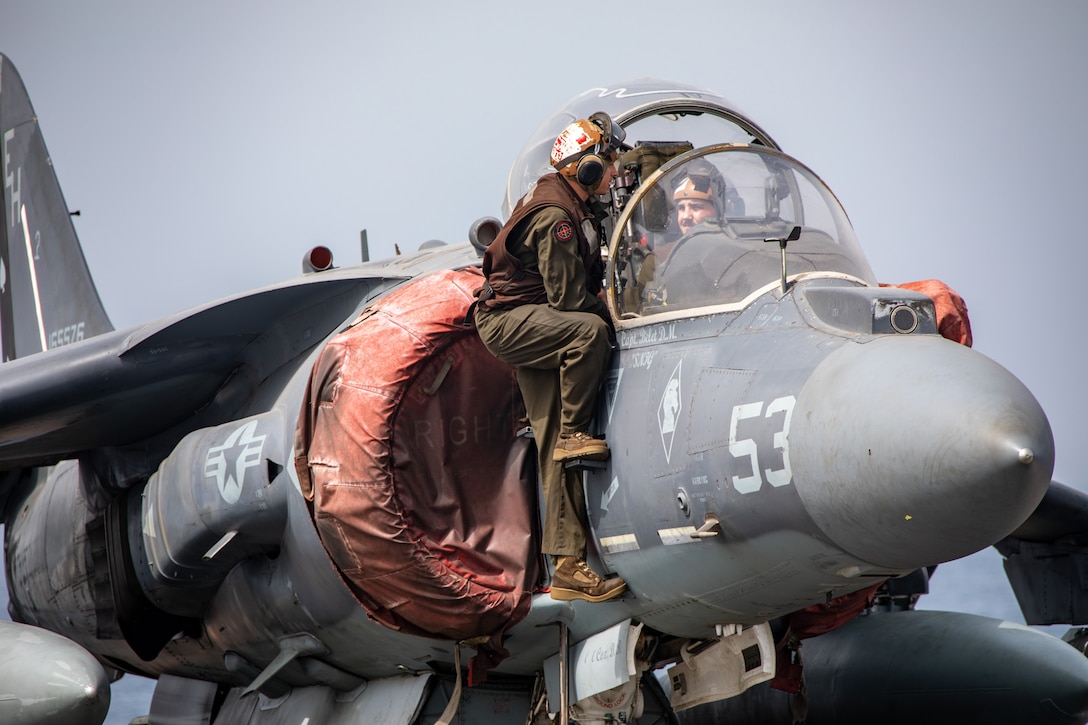 A Marine who has climbed up the side of a jet talks to another Marine in the cockpit.