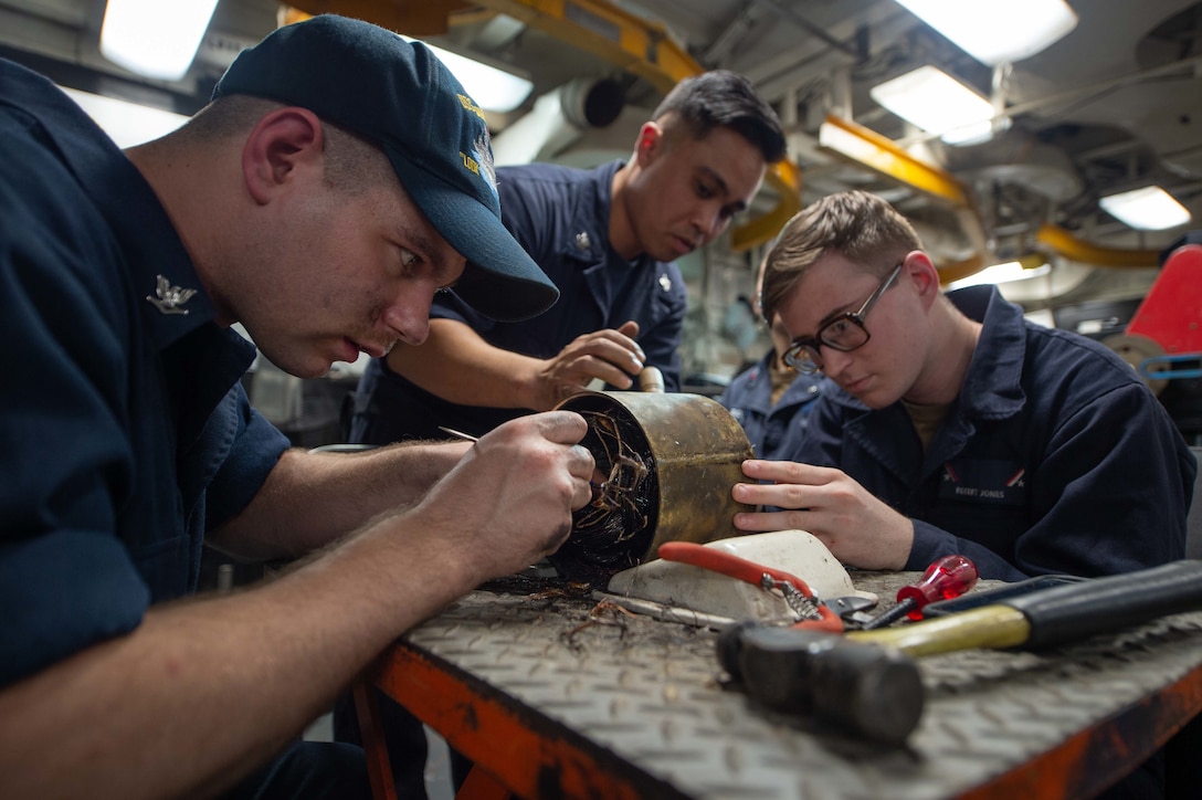 Three sailors around a workbench adjust the wiring on an electric motor as a hammer, screwdriver and pliers lay in the foreground.