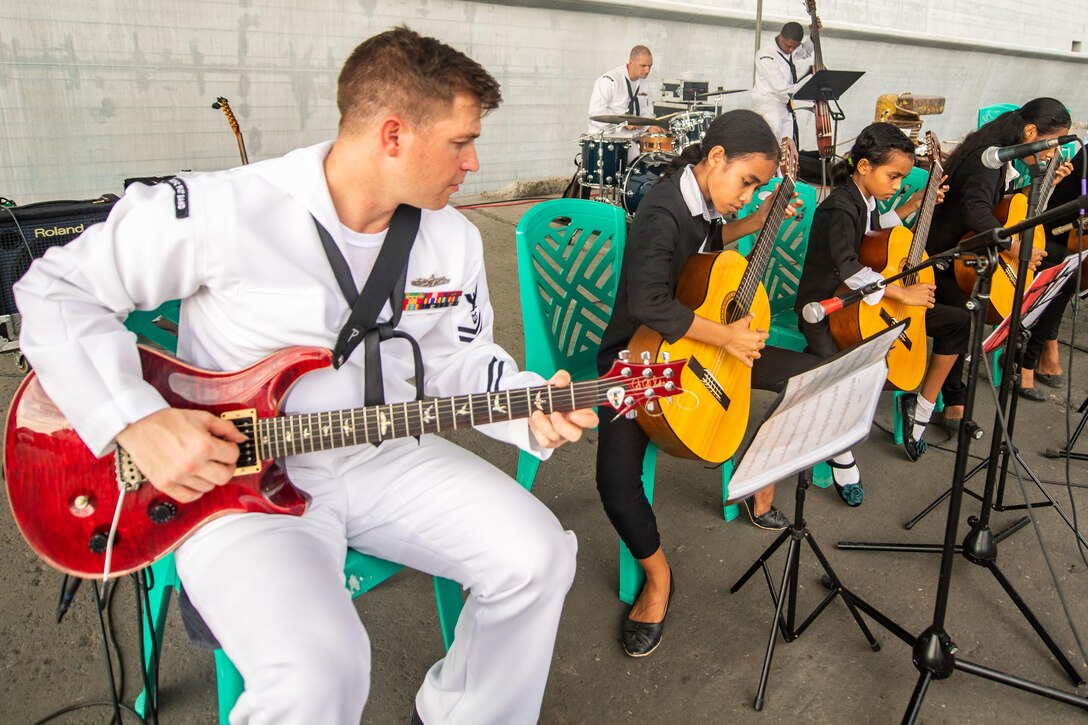 A sailor plays electric guitar  beside a group of students who are playing acoustic guitars.