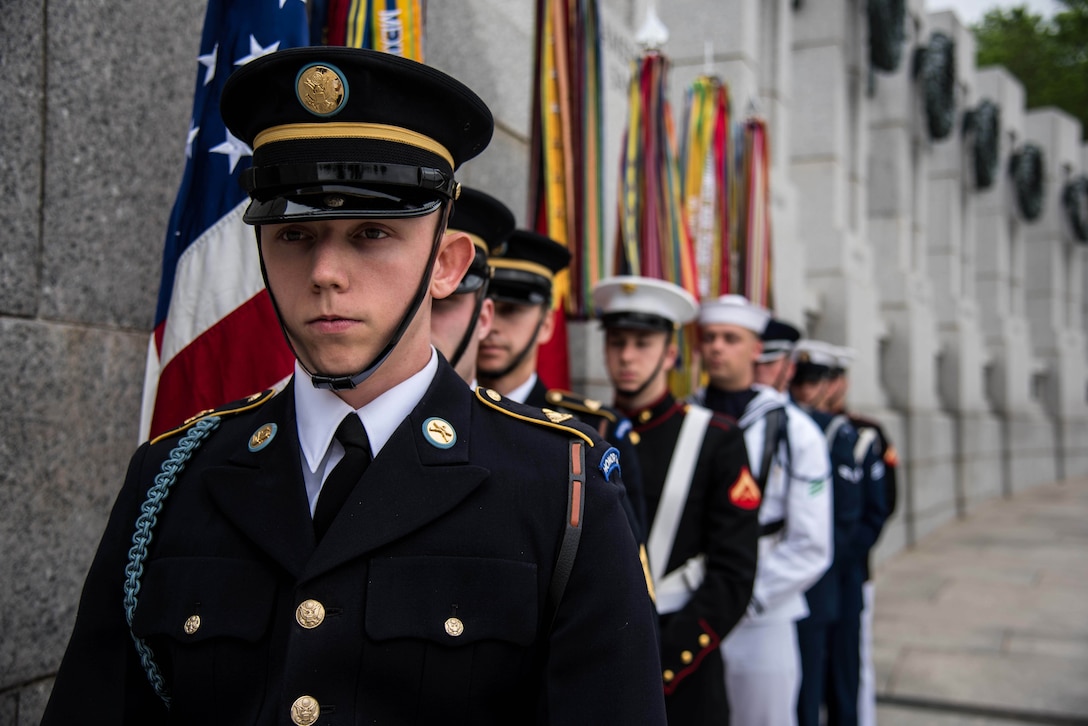 Eight service members in dress uniforms stand next to several flags at a memorial.