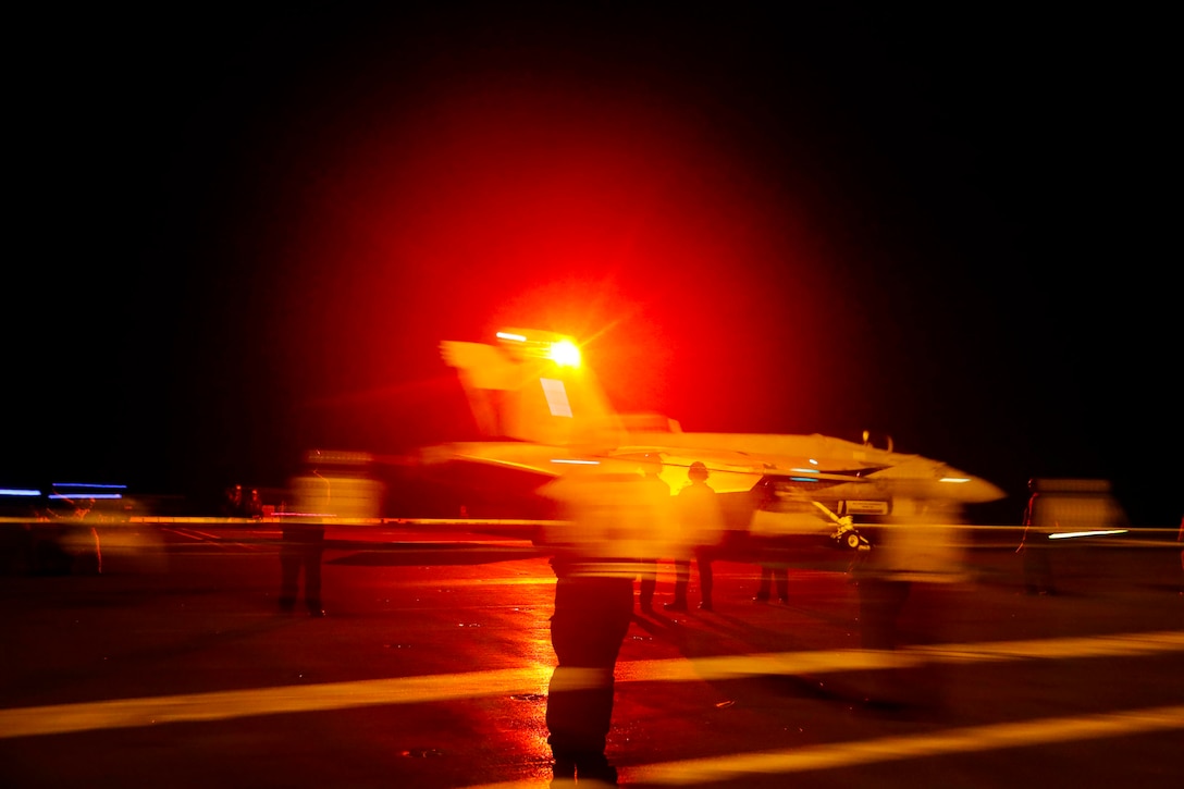 A military jet takes off from an aircraft carrier at night with a red light shining behind it.