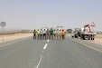 Members of the 1st Theater Sustainment Command and a Kuwait work crew stand for a photo in between two recently installed traffic signs during a traffic safety sign setup initiative conducted along routes in the vicinity of Camp Buehring Kuwait, May 11, 2019.