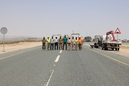 Members of the 1st Theater Sustainment Command and a Kuwait work crew stand for a photo in between two recently installed traffic signs during a traffic safety sign setup initiative conducted along routes in the vicinity of Camp Buehring Kuwait, May 11, 2019.