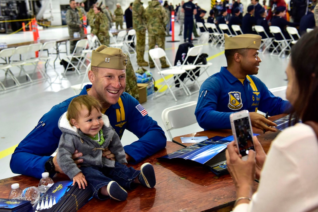 A sailor sits at a table holding a baby while posing for a photo.