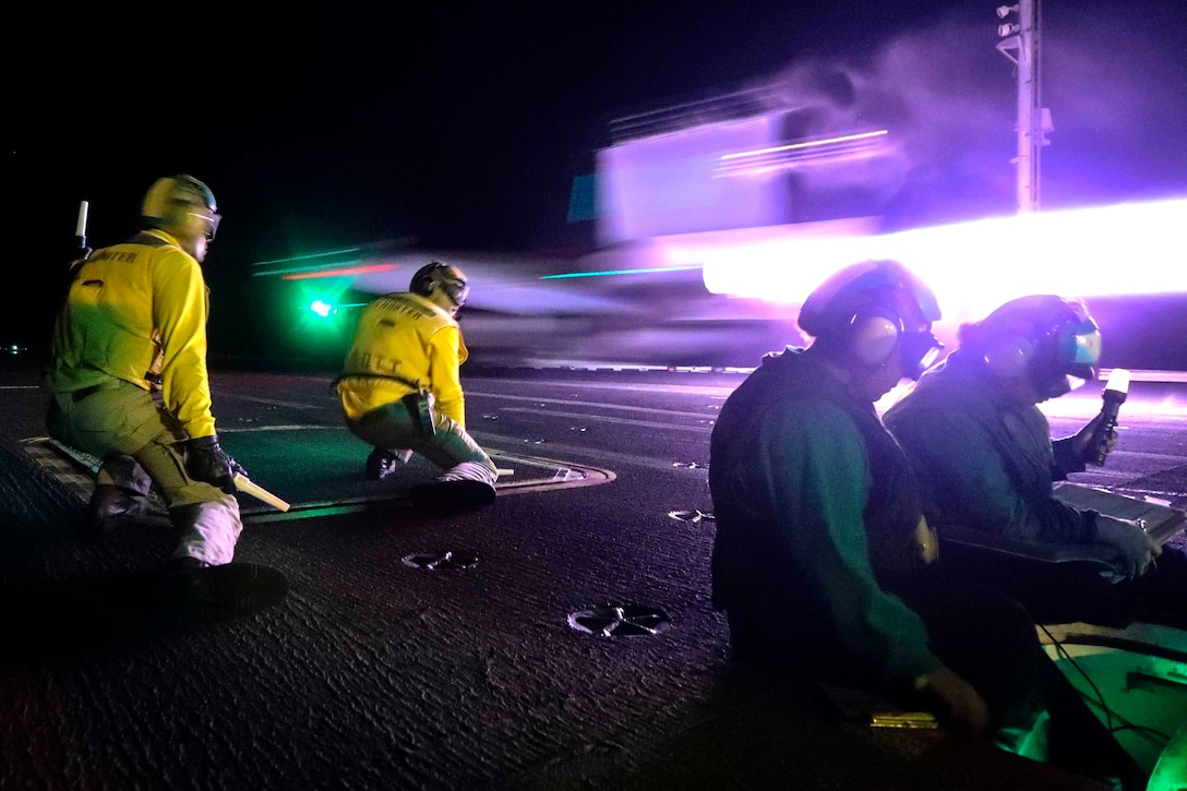 A military fighter jet takes off from an aircraft carrier at night as sailors kneel close by.