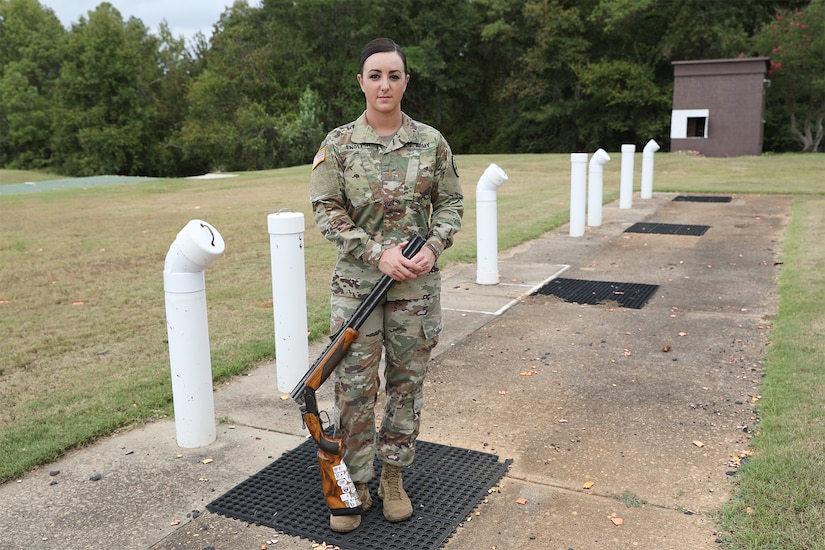 A female soldier in camouflage uniform poses with a shotgun at the firing line.