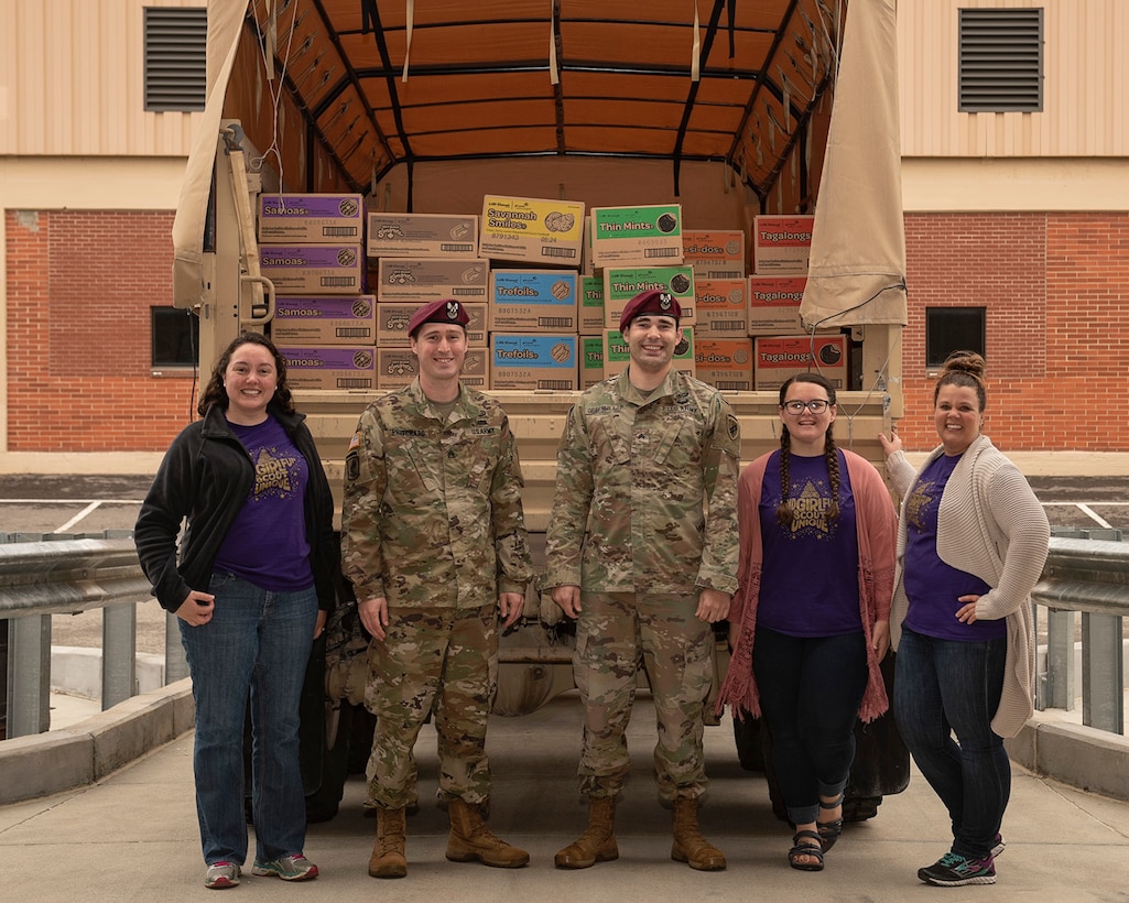 Three Girl Scouts and two military members stand in front of truck filled with cookies