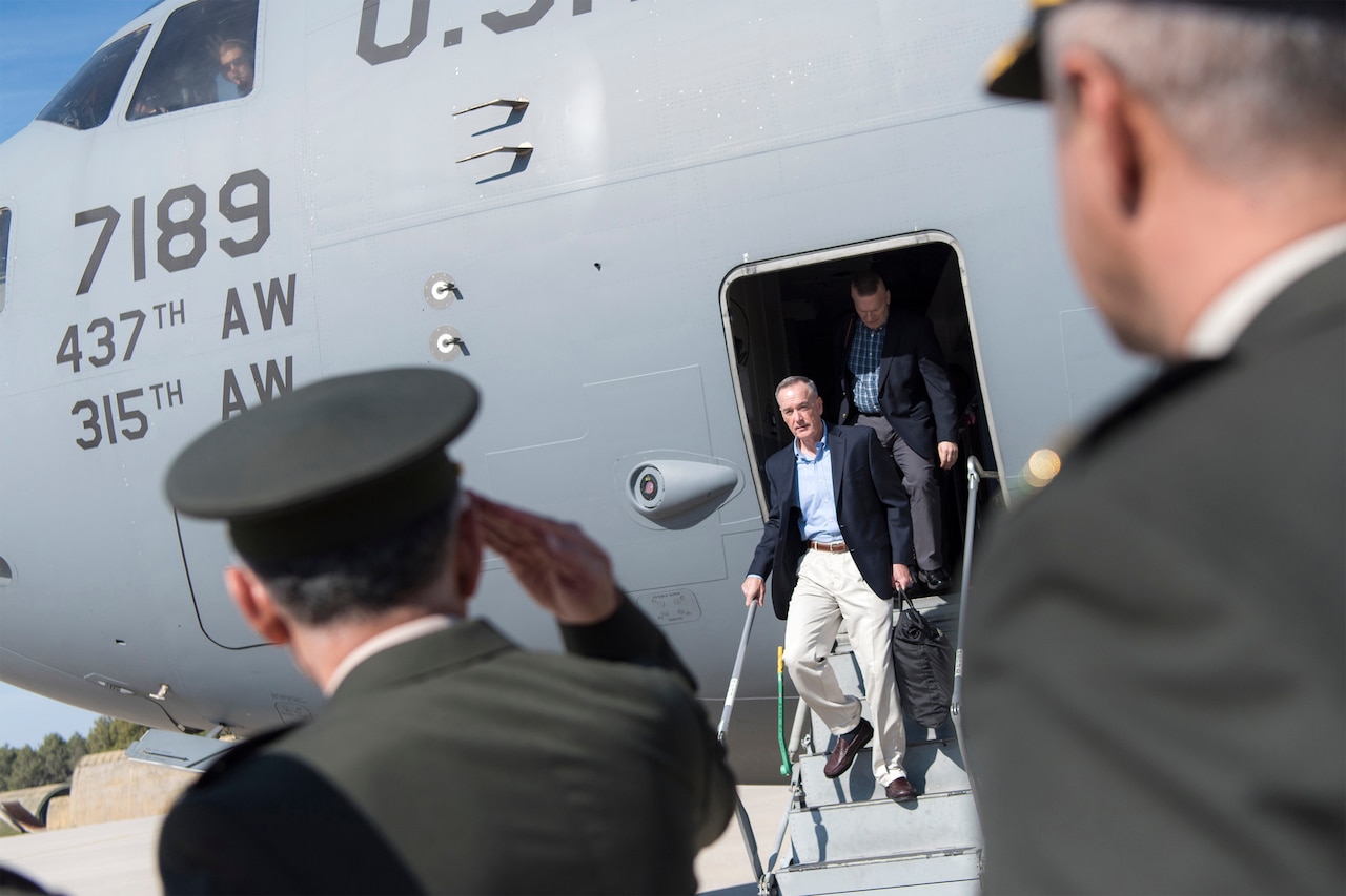 Men in uniform salute a man in civilian clothes exiting an airplane.