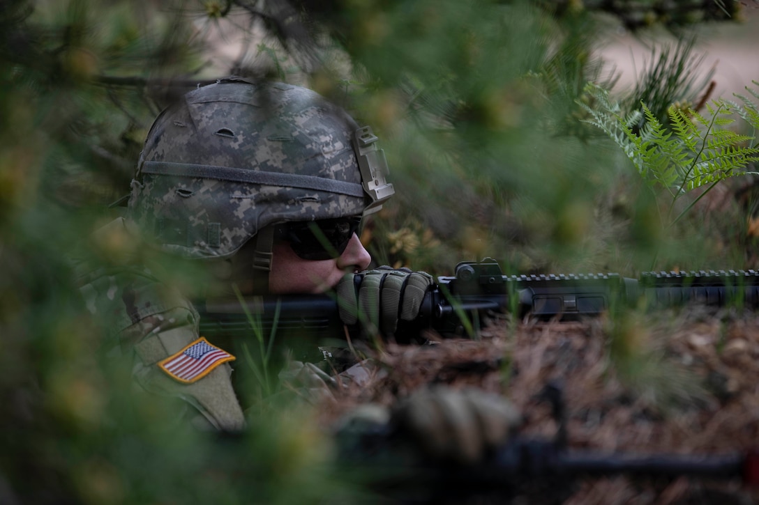 A soldier wearing a helmet and sunglasses crouches behind a berm and aims a rifle.