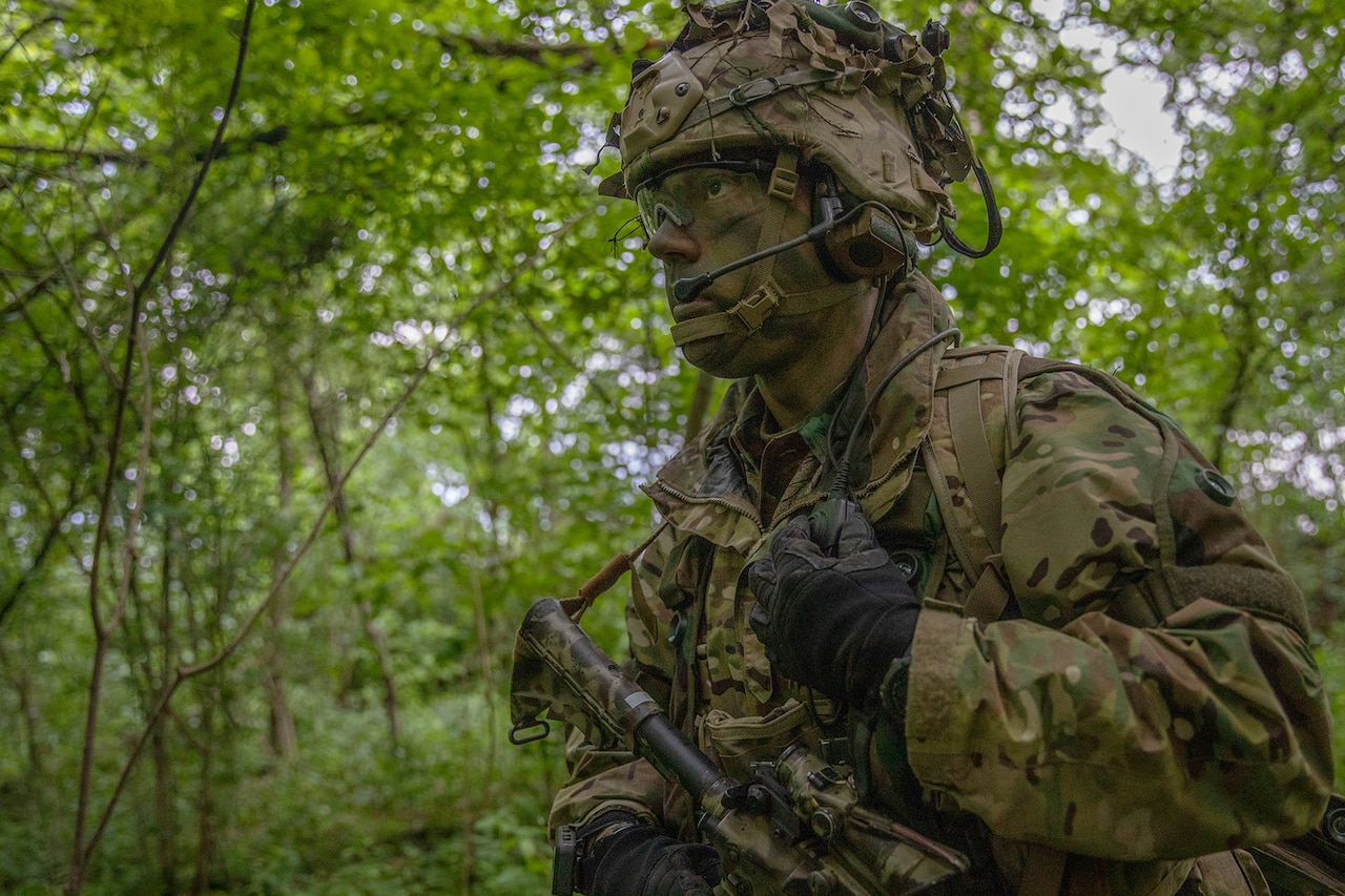Soldier in camouflage uniform and face paint walks in a wooded area.