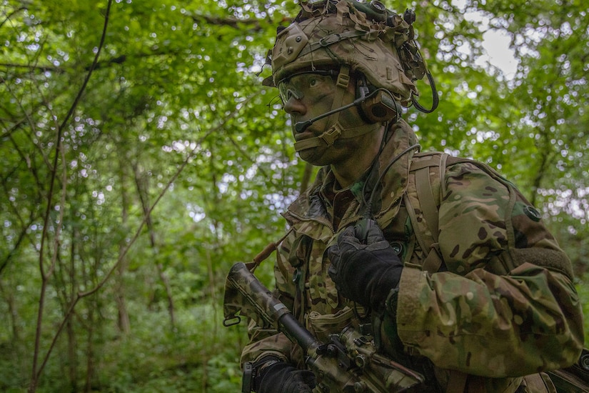 Soldier in camouflage uniform and face paint walks in a wooded area.