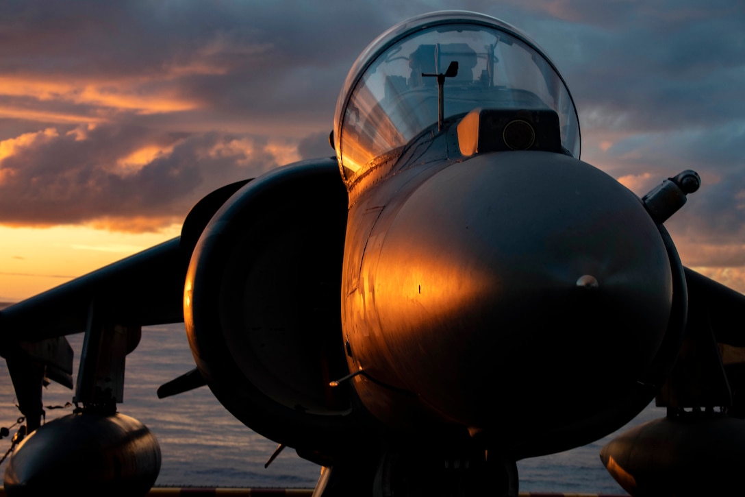 Sunlight reflects off a fighter jet as it waits on a flight deck with the ocean in the background.