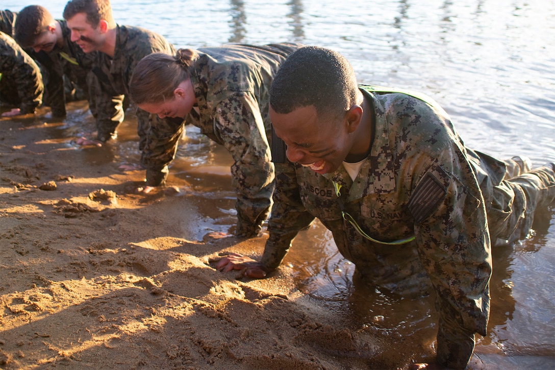 Navy midshipmen perform pushups along the water.