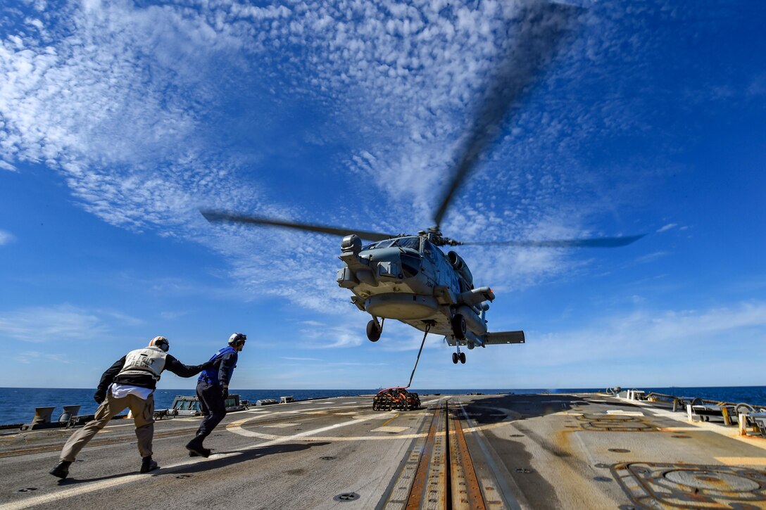 Two sailors walk toward a helicopter as it lands on a military ship.