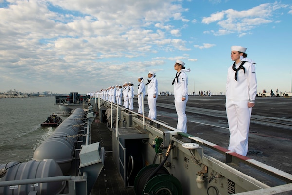 Sailors stand in line along the edge of the deck of a ship.