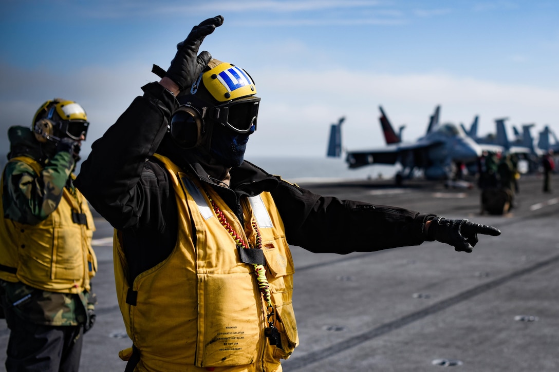 A sailor points his finger as he guides a military jet on an aircraft carrier.