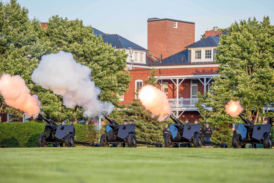 Soldiers stand by four firing cannons on a lawn with red brick buildings in the background.