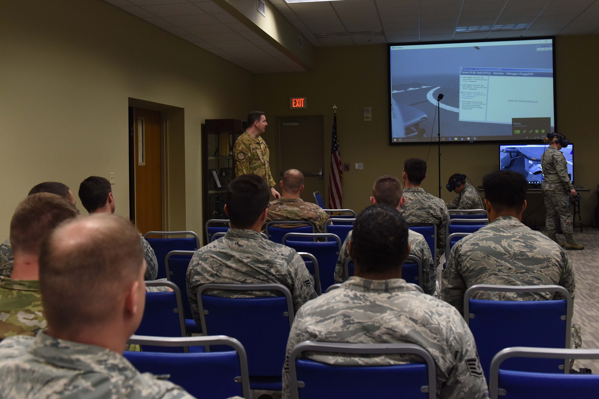 A man dressed in operational camouflage pattern speaks to a crowd of military service members in front of a large projected screen