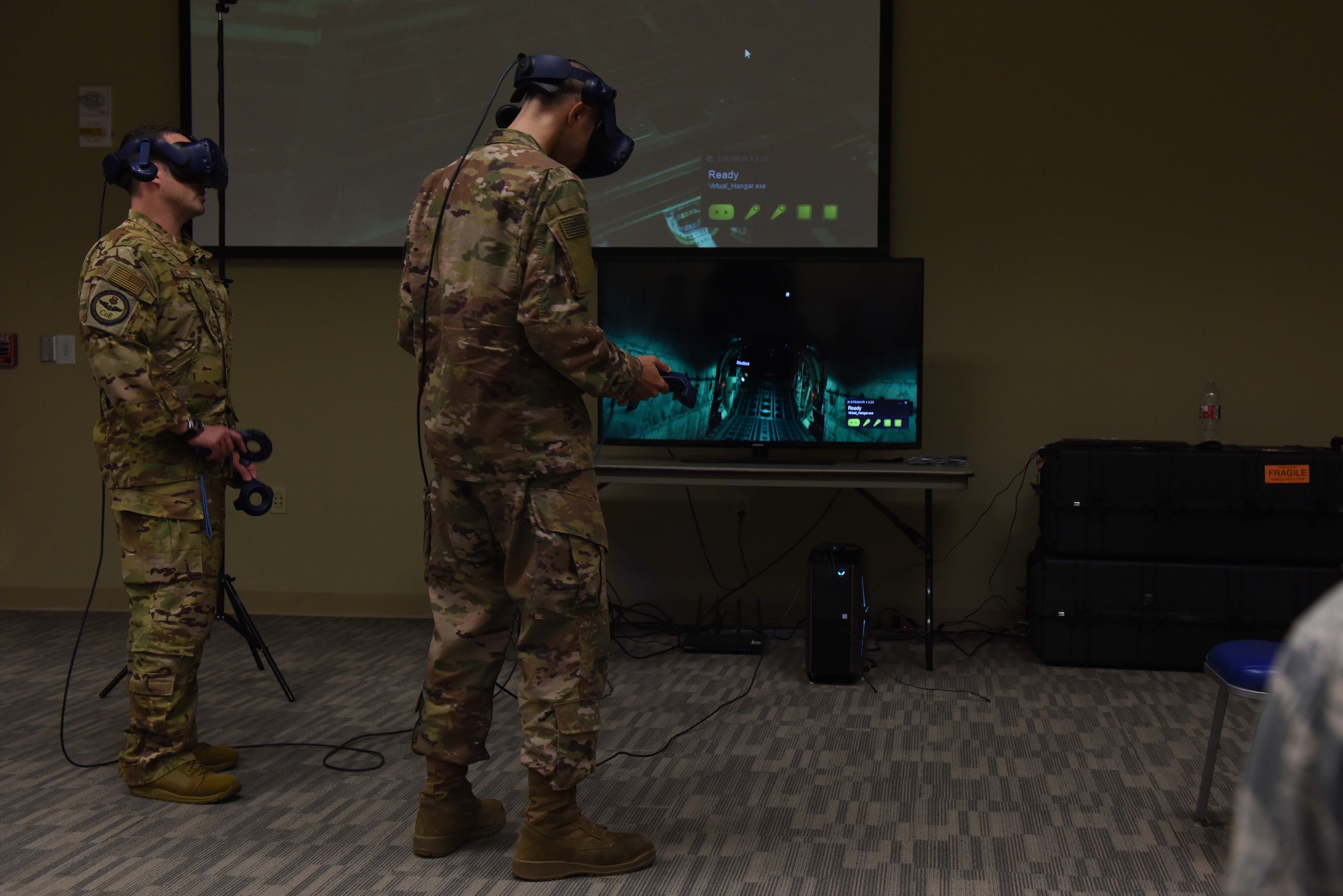 Two Airmen wearing occupational camouflage pattern uniform stand in front of a projector while using virtual reality gear