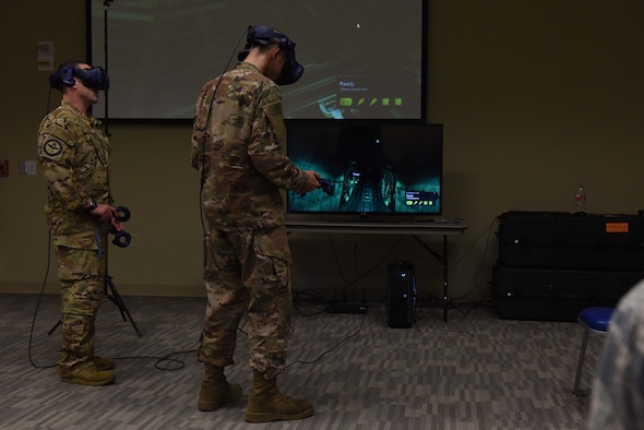 Two Airmen wearing occupational camouflage pattern uniform stand in front of a projector while using virtual reality gear