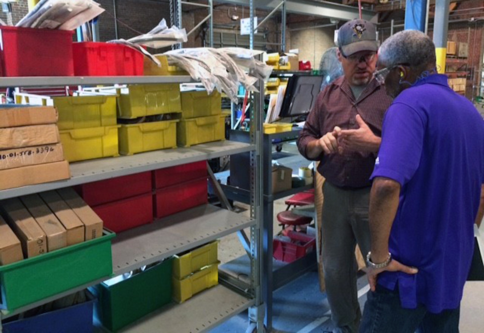 Two men discuss materials and parts in front of industrial shelving