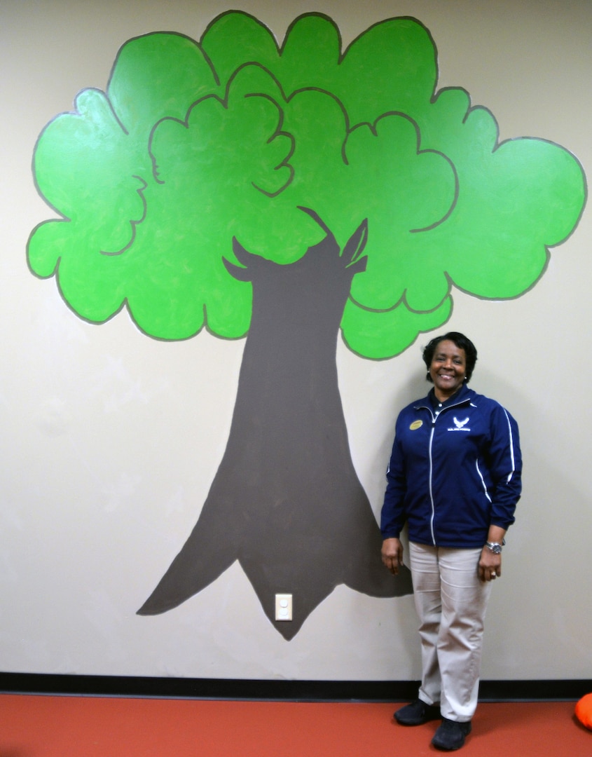 Feletia McLaurin, Medical Education and Training Campus Fitness Center fitness and sports manager, stands beside a mural of a tree she painted in the new Parent Child Area at the fitness that officially opens June 3. Located in a former workout room and storage space at the fitness center, the Parent Child Area has a play area and small table for children and includes a flat screen TV and DVD player for viewing children’s movies. In addition, it includes an area with new exercise equipment for parents to work out on while they watch over their children.