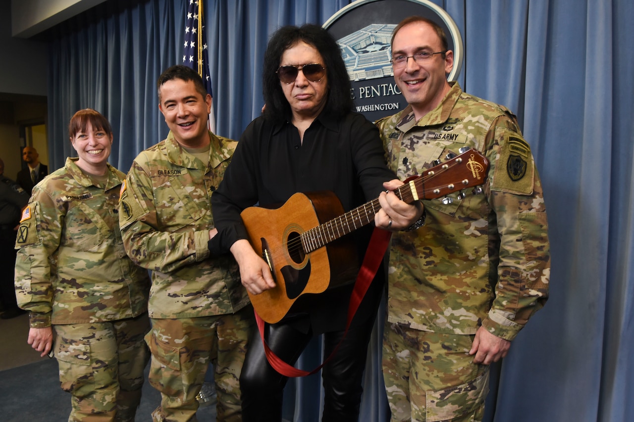 Famous musician Gene Simmons holds a guitar while standing next to three service members on the stage in the pentagon briefing room.