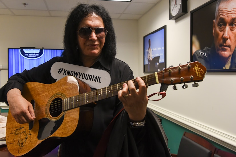 Famous musician Gene Simmons strums a guitar while wearing a #KNOWYOURMIL sign on his chest.