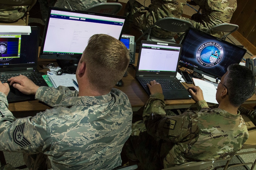 Two service members sit at a table with four computers.